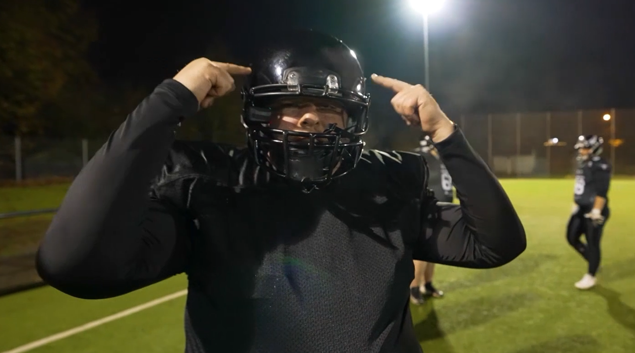 Reeze steht in Football-Schutzkleidung auf einem Sportplatz und zeigt mit beiden Händen auf seinen Kopf. (Quelle: BSI) Reeze steht in Football-Schutzkleidung auf einem Sportplatz und zeigt mit beiden Händen auf seinen Kopf.
