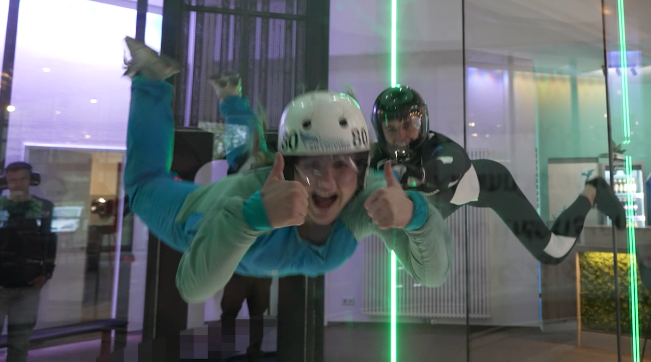 Ollimeee schwebt lachend neben einem Trainer beim Indoor-Skydiving in der Luft und streckt beide Daumen nach oben. (Quelle: BSI) Ollimeee schwebt lachend neben einem Trainer beim Indoor-Skydiving in der Luft und streckt beide Daumen nach oben.