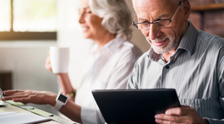 Älterer Mann mit Tablet und ältere Frau im Hintergrund mit Smartwatch am Laptop (Quelle: © zinkevych Fotolia.com) Älterer Mann mit Tablet und ältere Frau im Hintergrund mit Smartwatch am Laptop