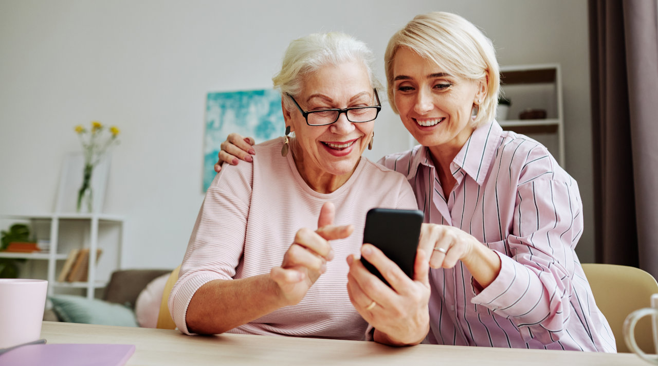 Zwei Frauen in einem Wohnzimmer schauen auf ein Mobiltelefon. (Quelle: © Seventyfour / Adobe Stock) Zwei Frauen in einem Wohnzimmer schauen auf ein Mobiltelefon.