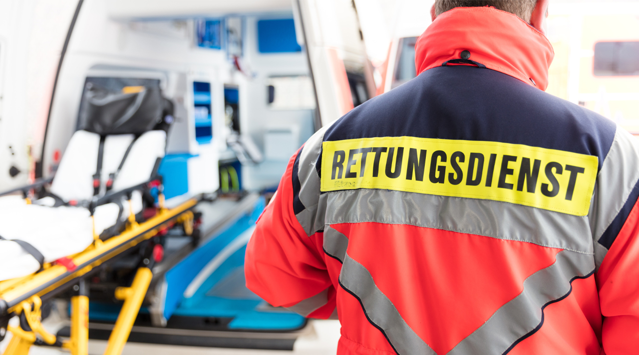 Ein Sanitäter mit einer Jacke auf der Rettungsdienst steht ist im Vordergrund des Bildes. Im Hintergrund steht ein offener Krankenwagen mit einer ausgezogenen Liege. (Quelle: © Tobias Seeliger / Adobe Stock ) Ein Sanitäter mit einer Jacke auf der Rettungsdienst steht ist im Vordergrund des Bildes. Im Hintergrund steht ein offener Krankenwagen mit einer ausgezogenen Liege.